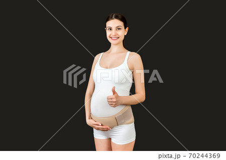 Portrait of young pregnant woman wearing bandage on her belly and showing thumb up sign at black background with copy space. Orthopedic abdominal support belt concept 70244369