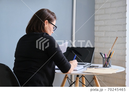 Mature woman sitting at desk with laptop, writing in business notebook Mature woman sitting at desk with laptop, writing in business notebook 70245465