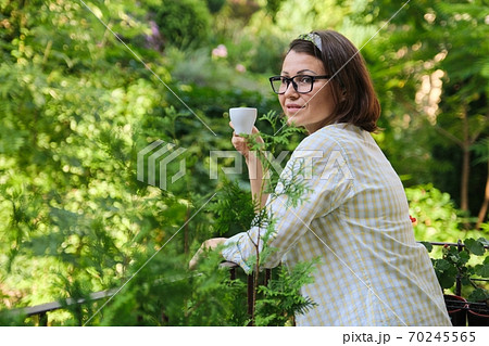 Mature woman standing on open balcony, female female with cup of coffee alone 70245565