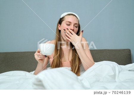 Sleepy yawning young woman in bed with cup of tea Sleepy yawning young woman in bed with cup of tea 70246139