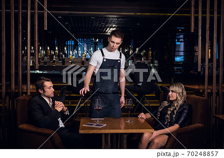 an experienced waiter in an apron pours red wine into glasses for a young blonde and a handsome man 70248857