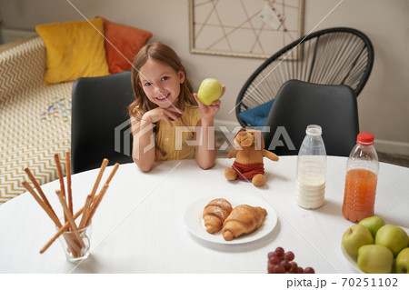Happy caucasian girl sitting at the table at home, holding green apple and smiling at camera, having breakfast at home Happy caucasian girl sitting at the table at home, holding green apple and smiling at camera, having breakfast at home 70251102