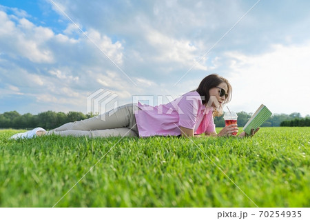 Mature woman reading book, female lying on green grass with drink 70254935