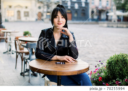 Happy young mixed raced woman sitting at cafe at the table and looking at camera. Chinese lady in black leather jacket spending free time at outdoor cafe. Old ancient buildings on the background 70255994