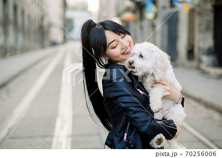 A young dark haired Asian woman, dressed in black leather jacket, stands on street in old European city street, holding and embracing white little dog. Horizontal urban shot, copy space 70256006