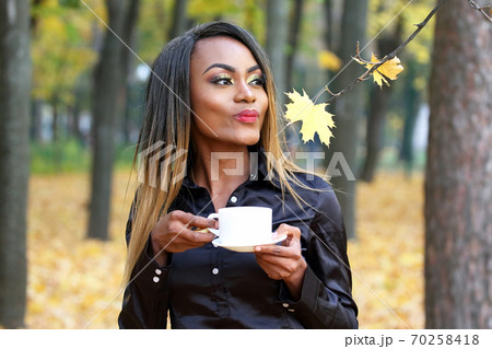 Beautiful young African woman drinking coffee from a white Cup on the background of autumn leaves  70258418