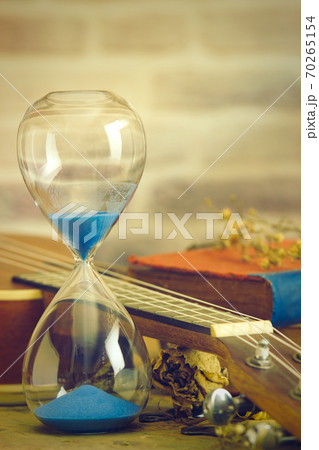 A vintage hourglass and ukulele with an old book and brass pen on a wooden table and brick background in the morning. Closeup and copy space. The concept of memories or things in the past. A vintage hourglass and ukulele with an old book and brass pen on a wooden table and brick background in the morning. Closeup and copy space. The concept of memories or things in the past. 70265154