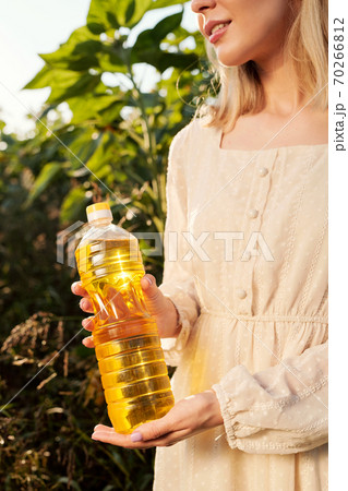 Blond young woman in stylish white dress holding bottle of sunflower oil Blond young woman in stylish white dress holding bottle of sunflower oil 70266812