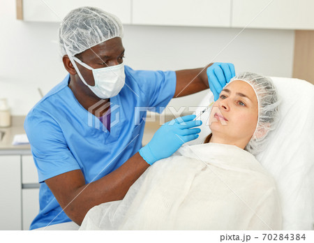 Doctor in mask is doing injection to woman patient in skin of face in estetic clinic 70284384