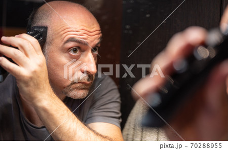 A middle-aged Caucasian man cuts his hair in the bathroom. While placing the clipper on your head check with your eyes in the mirror. A middle-aged Caucasian man cuts his hair in the bathroom. While placing the clipper on your head check with your eyes in the mirror. 70288955