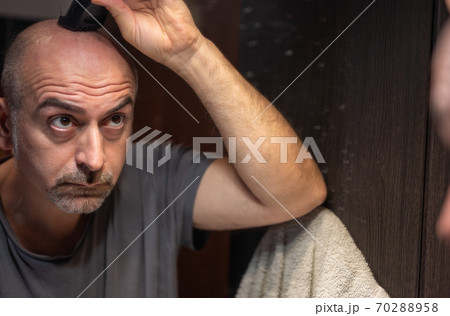 A middle-aged Caucasian man cuts his hair in the bathroom. While placing the clipper on your head check with your eyes in the mirror. 70288958