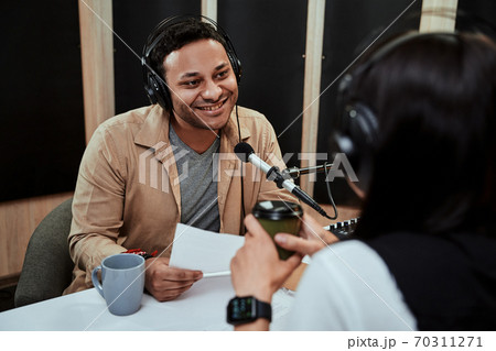 Portrait of young male radio host going live on air, talking with female guest, holding a script paper while sitting in studio 70311271