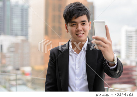 Portrait of happy young Asian businessman taking selfie in the city Portrait of happy young Asian businessman taking selfie in the city 70344532