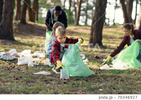 Purposeful boy picking up rubbish on plastic bag with volunteers of different ages in the park. Anti pollution concept. 70346927