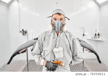 Portrait of professional auto painter. Worker painting parts of the car in special painting chamber, wearing costume and protective gear. Car service station. 70350611