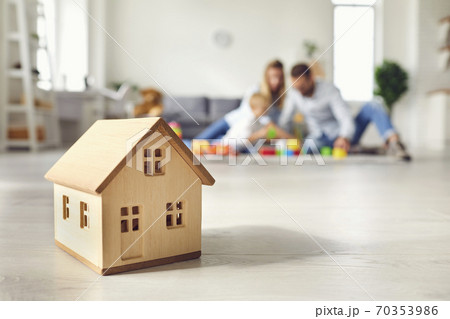 Little wooden house on floor of cozy room with happy family playing in background Little wooden house on floor of cozy room with happy family playing in background 70353986