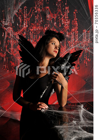 Beautiful young brunette woman in black angel costume with wings over spooky red background posing by piano with rose bouquet. Halloween party outfit 70356336