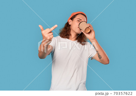 Freestyle. Curly-haired guy wearing hat standing isolated on blue drinking coffee showing middle finger cool 70357622