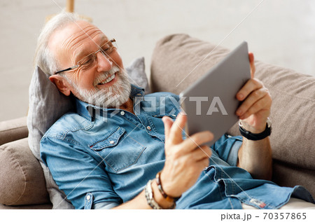 Cheerful aged man using tablet on couch. Cheerful aged man using tablet on couch. 70357865