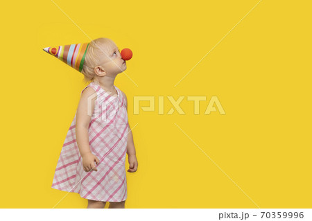 Toddler girl wearing party hat and red clown nose and looks up. Portrait on yellow background. Copy space. Toddler girl wearing party hat and red clown nose and looks up. Portrait on yellow background. Copy space. 70359996