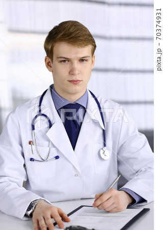 Blond male doctor sitting and working with clipboard in clinic at his working place, close-up. Young physician at work. Perfect medical service, medicine concept 70374931