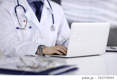 Unknown male doctor sitting and working with laptop in clinic at his working place, close-up. Young physician at work. Perfect medical service, medicine concept Unknown male doctor sitting and working with laptop in clinic at his working place, close-up. Young physician at work. Perfect medical service, medicine concept 70374944