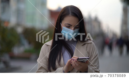 Portrait woman in protective medical mask walks down to the street uses phone texts scrolls surfs the internet search news of second wave covid-19 coronavirus virus protection pandemic Portrait woman in protective medical mask walks down to the street uses phone texts scrolls surfs the internet search news of second wave covid-19 coronavirus virus protection pandemic 70378089