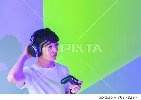 【eスポーツのイメージ】紫と緑の照明のもとコントローラを片手にエキサイトしている男性ゲーマー 70378237