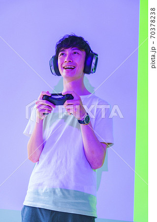 【eスポーツのイメージ】紫と緑の照明のもとコントローラを片手にエキサイトしている男性ゲーマー 70378238