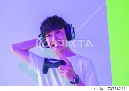 【eスポーツのイメージ】紫と緑の照明のもとコントローラを片手にエキサイトしている男性ゲーマー 70378241