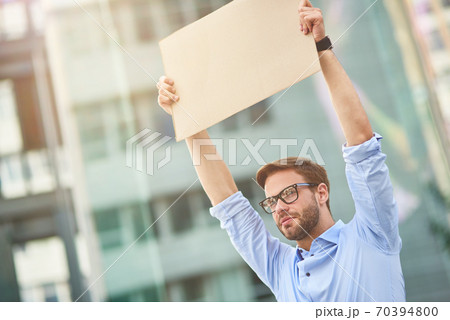 Protesting outdoors. Portrait of a young male activist wearing blue shirt and eyeglasses holding empty sign board over the head while standing outdoors Protesting outdoors. Portrait of a young male activist wearing blue shirt and eyeglasses holding empty sign board over the head while standing outdoors 70394800