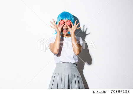 Image of happy asian girl in blue wig holding macarons over eyes and smiling coquettish, standing over white background Image of happy asian girl in blue wig holding macarons over eyes and smiling coquettish, standing over white background 70397138