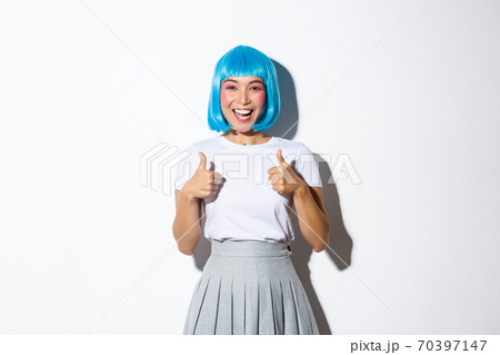 Cheerful cute asian girl in blue wig looking satisfied, smiling and showing thumbs-up, wearing halloween outfit, standing over white background 70397147