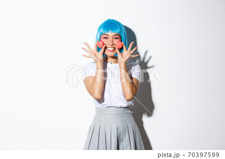 Image of beautiful smiling asian girl looking silly, showing macaroons, wearing blue anime wig, standing over white background Image of beautiful smiling asian girl looking silly, showing macaroons, wearing blue anime wig, standing over white background 70397599