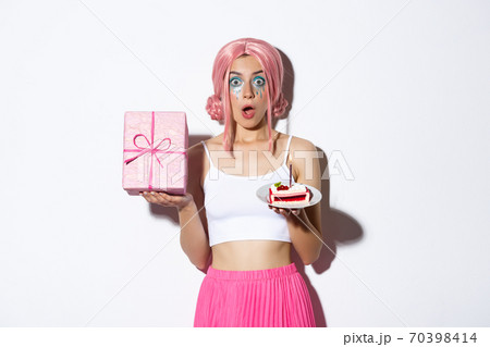 Portrait of surprised beautiful girl in pink wig, receive birthday gift, holding b-day cake and smiling happy, standing over white background 70398414