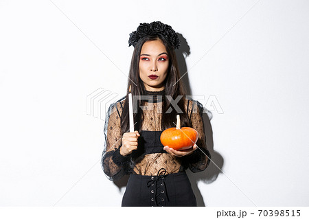 Image of asian woman in wicked witch costume, looking left serious, holding lit candle and pumpkin, celebrating halloween Image of asian woman in wicked witch costume, looking left serious, holding lit candle and pumpkin, celebrating halloween 70398515