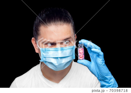 Doctor virologist in medical mask and sterile gloves raising an eyebrow contemptuously looking at a glass bottle filled with pills for treating coronavirus, isolated on a black background. 70398718