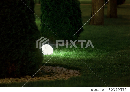 illumination backyard light garden with lantern electric lamp with a round diffuser in the green grass with thuja bushes in a park with landscaping, closeup night scene nobody. 70399535