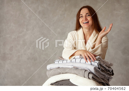 Young woman with beautiful smile in warm beige robe is leaning on stack of folded bed sheets 70404562