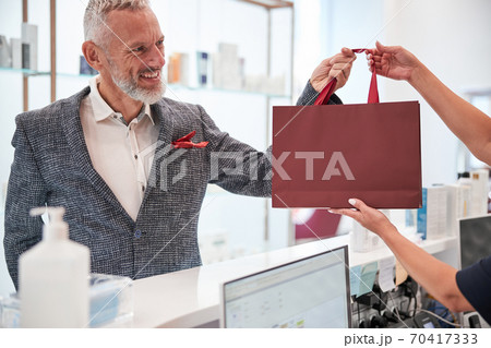 Cheerful elderly man taking a red bag from a shop assistant Cheerful elderly man taking a red bag from a shop assistant 70417333