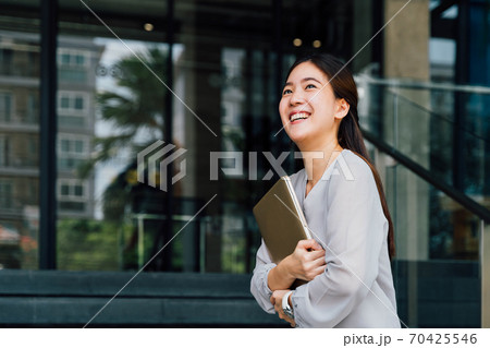 One successful and happy young adult business woman holding a laptop with smile. Professional female worker standing in corporate modern office and window in casual gray look. - With copy space One successful and happy young adult business woman holding a laptop with smile. Professional female worker standing in corporate modern office and window in casual gray look. - With copy space 70425546