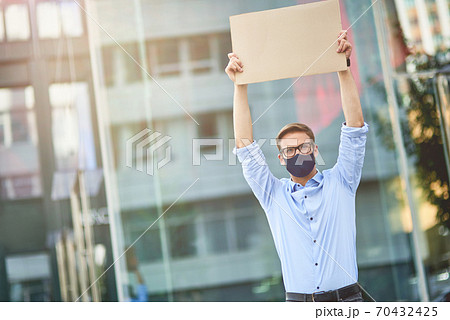 Young caucasian man, male activist wearing black protective mask holding empty sign board over the head while standing on city street. Protest during coronavirus outbreak 70432425