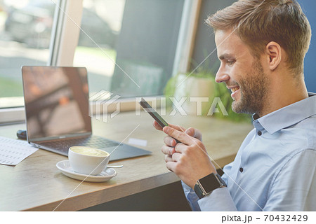 Chatting with friends. Side view of a freelancer or businessman using his smartphone and smiling while working remotely in cafe, drinking coffee Chatting with friends. Side view of a freelancer or businessman using his smartphone and smiling while working remotely in cafe, drinking coffee 70432429