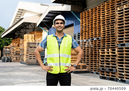 Portrait of young Indian worker working in logistic industry outdoor in front of factory warehouse. Smiling happy man with hard hat looking at camera hands on hip at depot 70440509