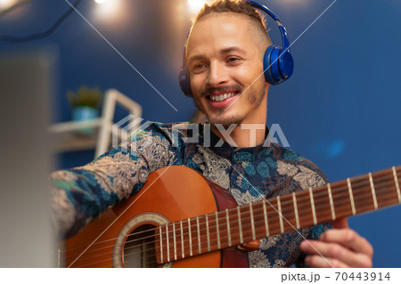Young man sitting in his room and watching guitar tutorial on laptop Young man sitting in his room and watching guitar tutorial on laptop 70443914