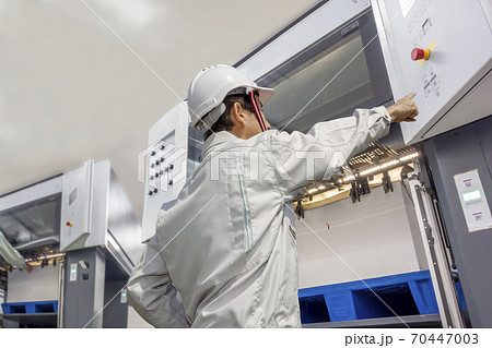 印刷工場で枚葉オフセット印刷機を操作する作業着を着た男性オペレーターの後ろ姿の上半身。 印刷工場で枚葉オフセット印刷機を操作する作業着を着た男性オペレーターの後ろ姿の上半身。 70447003