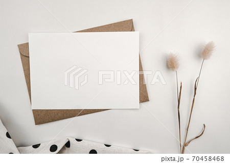 Blank white paper on brown paper envelope with Rabbit tail dry flower and White cloth with black dots. Mock-up of horizontal blank greeting card. Top view of Craft envelope on white background. 70458468