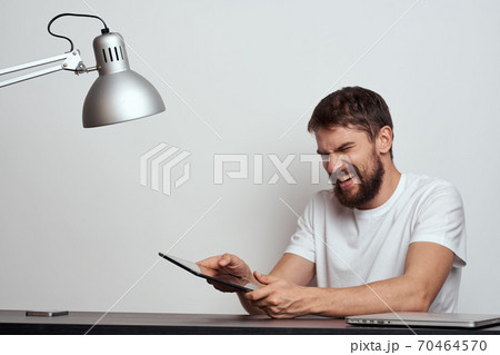 A man with a tablet at the table gestures with his hands on a light background and an iron lamp A man with a tablet at the table gestures with his hands on a light background and an iron lamp 70464570