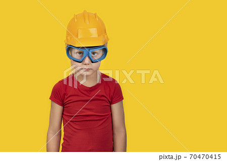 Blond boy in orange protective helmet and safety glasses on yellow background. Copy space 70470415