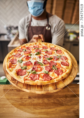 Male pizzeria worker in medical mask holding pepperoni pizza Male pizzeria worker in medical mask holding pepperoni pizza 70475419